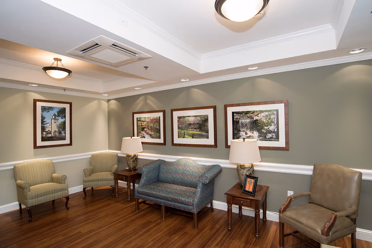 A cozy sitting area in a senior living facility with two striped armchairs, a patterned loveseat, and a leather armchair arranged around two wooden side tables with decorative lamps. The walls are painted light green and adorned with framed landscape photographs. The floor is wood, and the ceiling has recessed lighting and a central light fixture.