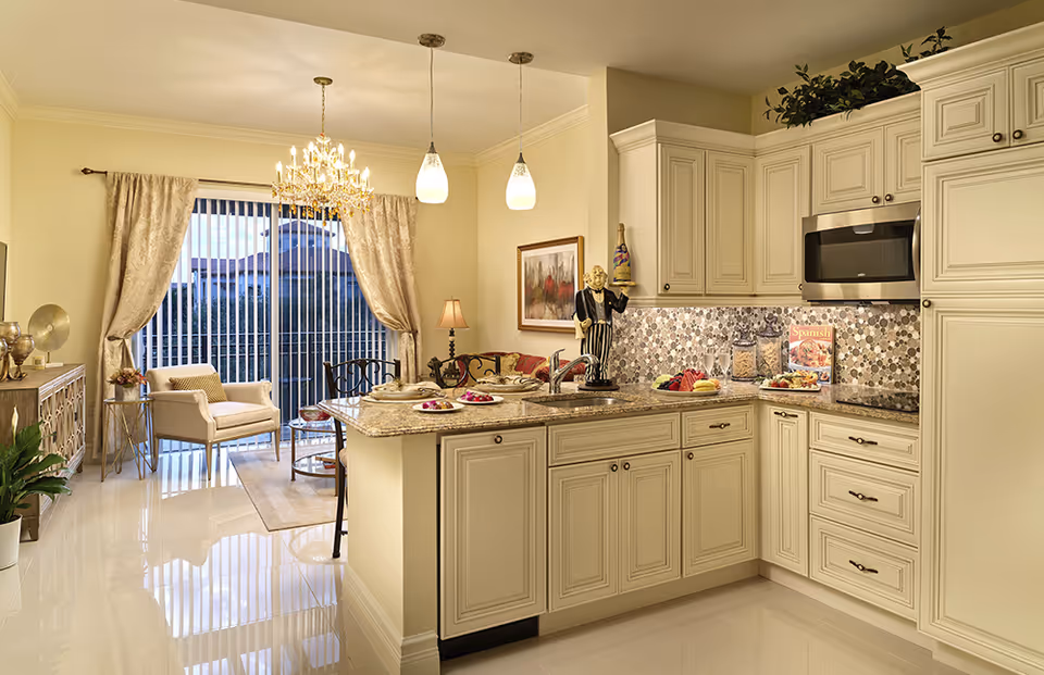 A bright and elegant kitchen and living area with cream-colored cabinetry, granite countertops, and a mosaic tile backsplash. The kitchen island has a sink and is set with plates and glasses. The living area features a chandelier, beige curtains, a comfortable armchair, a small table, and a decorative sideboard with plants and ornaments. Large sliding glass doors with vertical blinds lead to an outdoor view.