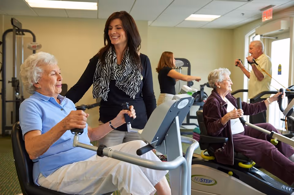 Two elderly women exercising on seated fitness machines in a gym with a younger woman assisting one of them. In the background, another elderly man and woman are also using exercise equipment in a well-lit room with large windows.