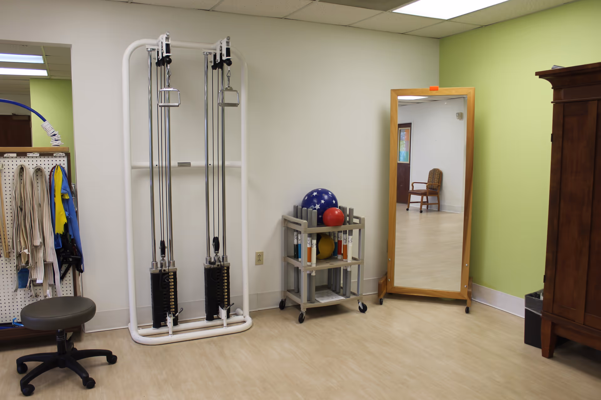 Interior therapy room with a resistance machine, cart of exercise balls, full-length mirror, stool and storage cabinet.