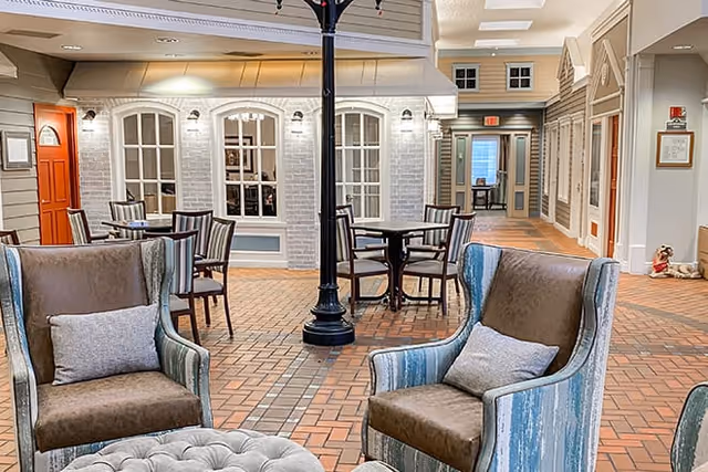 Indoor common area with armchairs, round tables, a central lamppost and storefront-style interior façades on a brick floor.