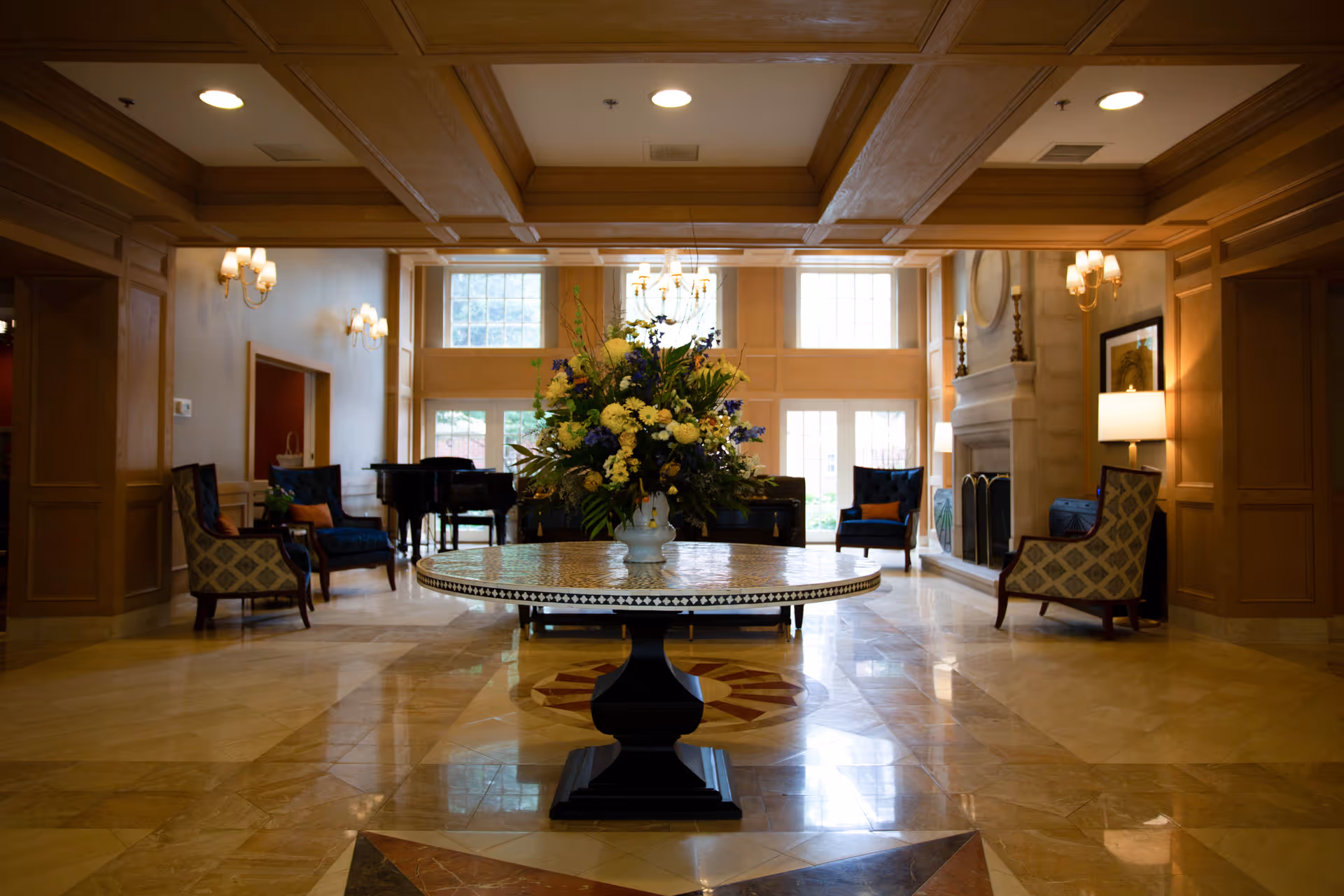 A spacious and elegant living room area with a large round table in the center holding a floral arrangement. The room features high ceilings with wooden beams, large windows allowing natural light, a fireplace on the right, and several upholstered chairs arranged around the room. The floor is made of polished stone tiles with decorative patterns.