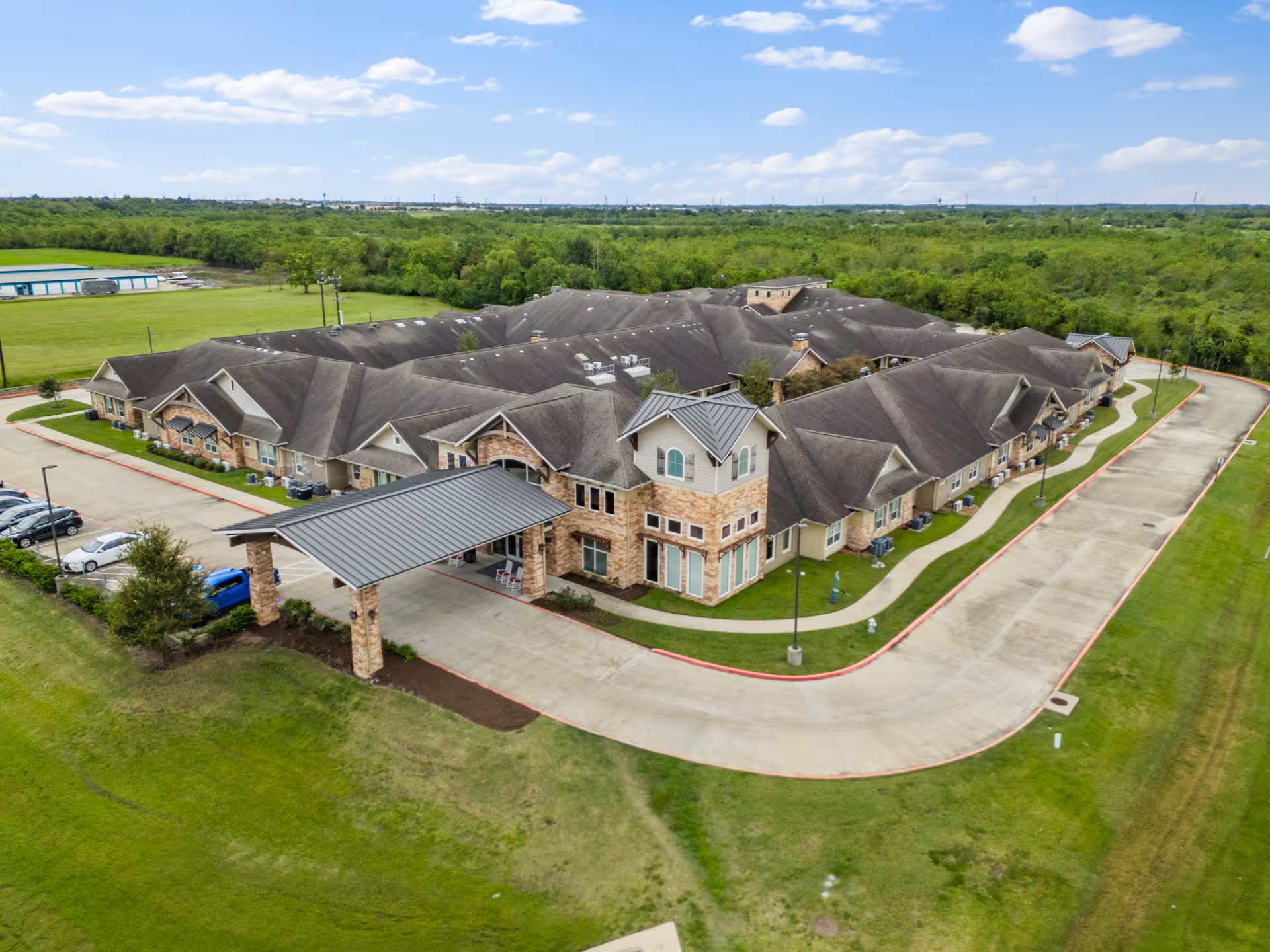 Aerial view of Discovery Village Clear Lake, a large senior living facility with a covered entrance, surrounded by green lawns and trees under a partly cloudy sky.