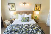 A neatly made bed with a tufted beige headboard, white pillows, and decorative floral pillows. The bedspread has a floral pattern in shades of gray, white, and yellow. On either side of the bed are wooden nightstands with lamps and decorative items. Two framed botanical prints hang on the wall above the headboard.