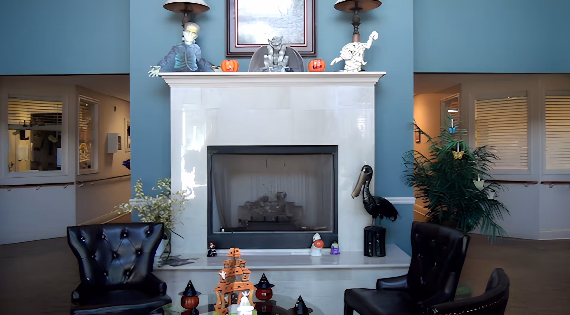 Interior seating area with a central marble fireplace decorated with Halloween figures, two black armchairs and potted plants.