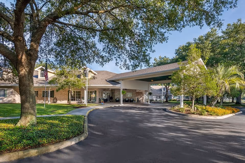 Covered main entrance and circular driveway of a senior living facility with landscaped islands and large trees.