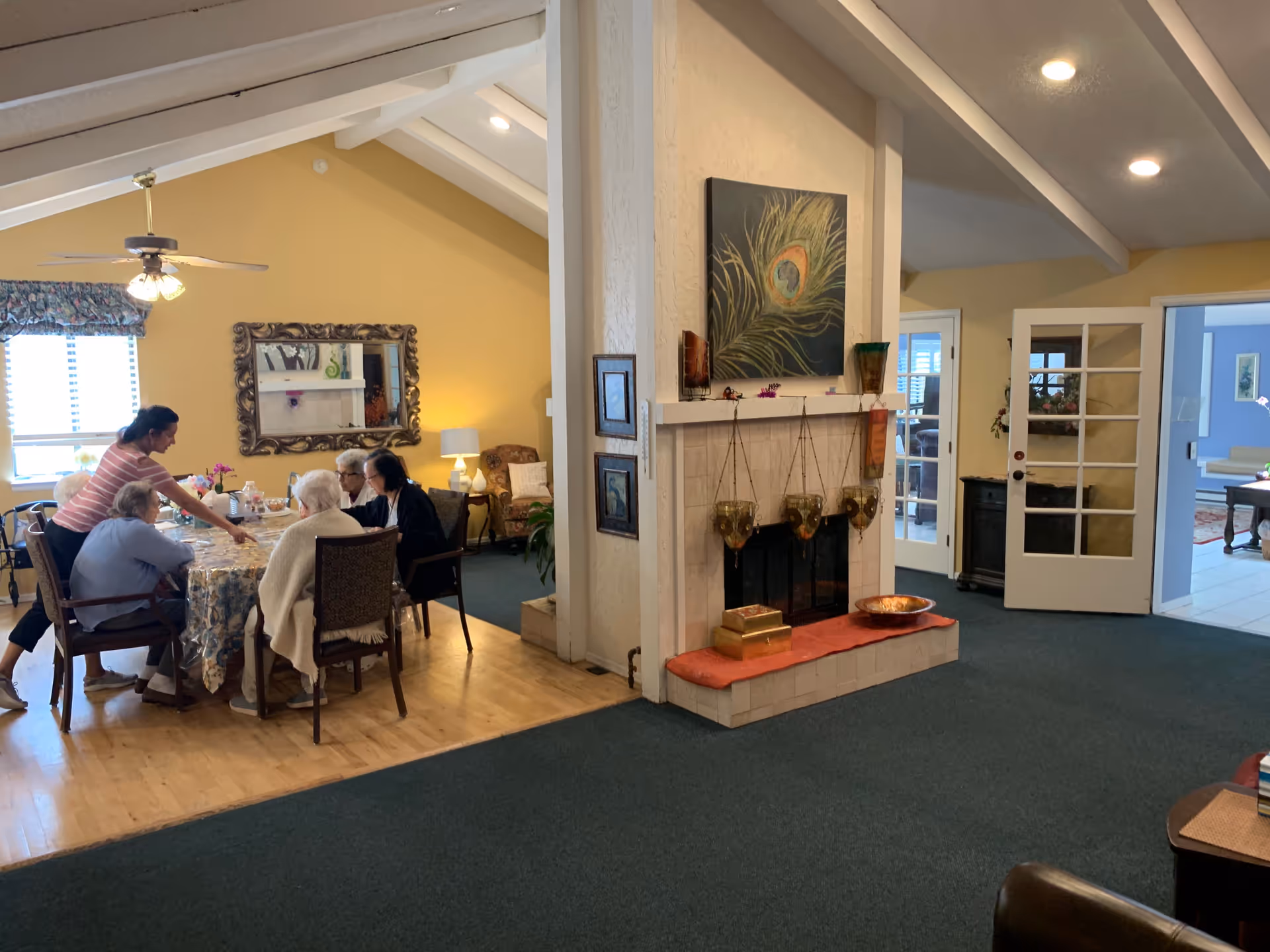 A cozy interior of Serenity House IV showing a group of elderly people seated around a dining table with a caregiver assisting them. The room has yellow walls, a ceiling fan, a large decorative mirror, and a fireplace with artwork above it. French doors lead to another room with blue walls.