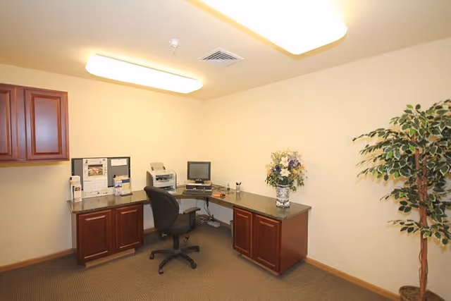 Small interior office with an L-shaped desk, swivel chair, computer and printer, cabinets, and a potted plant.