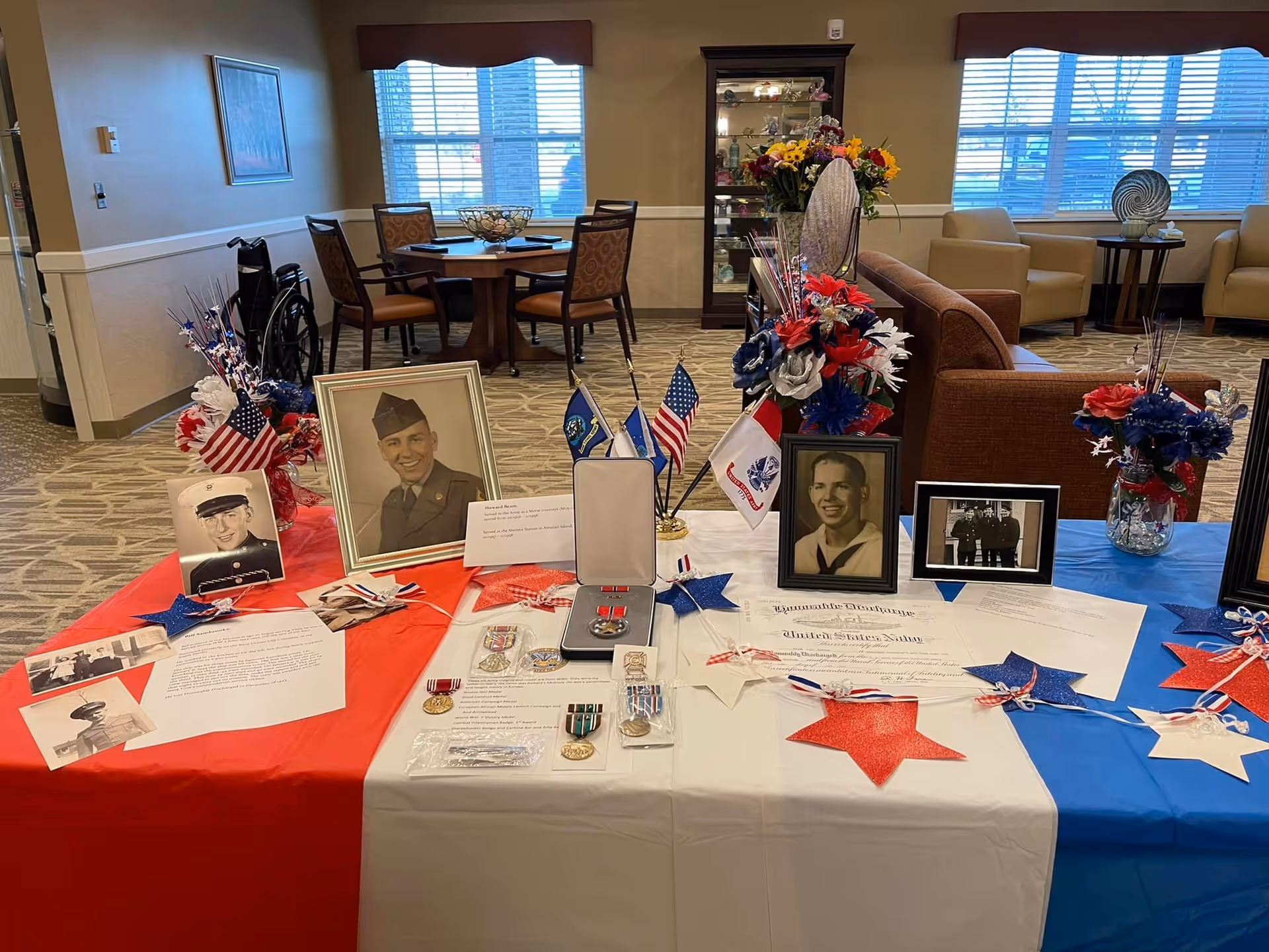 A patriotic display table covered with red, white, and blue cloths featuring framed black-and-white photographs of military personnel, medals, certificates, and decorative stars. The background shows a seating area with chairs, a table, and a cabinet with glass doors in a well-lit room.