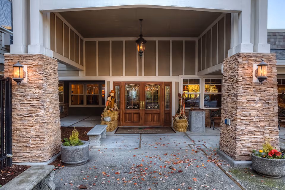 Entrance to a building with double wooden doors featuring decorative glass panels, flanked by stone pillars with wall-mounted lanterns. Seasonal decorations including scarecrows and hay bales are placed near the doors. There are concrete planters with plants and a stone bench near the entrance.