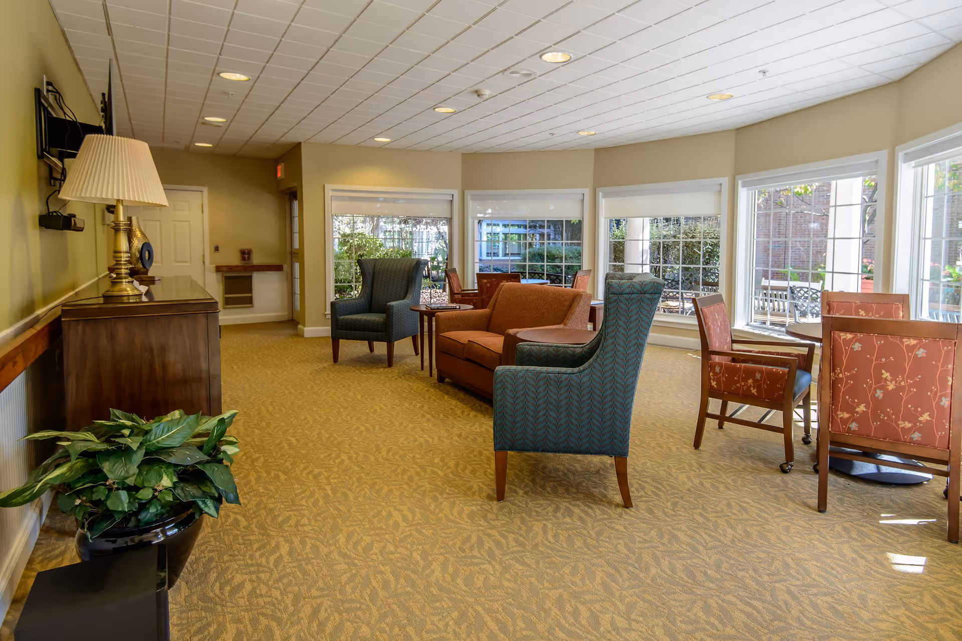 A bright and spacious senior living community lounge area with large windows letting in natural light. The room features a patterned carpet, two blue armchairs, a brown loveseat, and several tables with red cushioned chairs. A wooden sideboard with a lamp and decorative items is against one wall, and a potted plant is on the floor nearby.