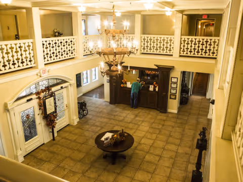View of a spacious lobby area in a senior living facility with tiled floors, a round table with decorative items in the center, a chandelier hanging from the ceiling, and a person standing at a reception desk. The lobby has a second-floor balcony with decorative railings and multiple doorways leading to other areas.