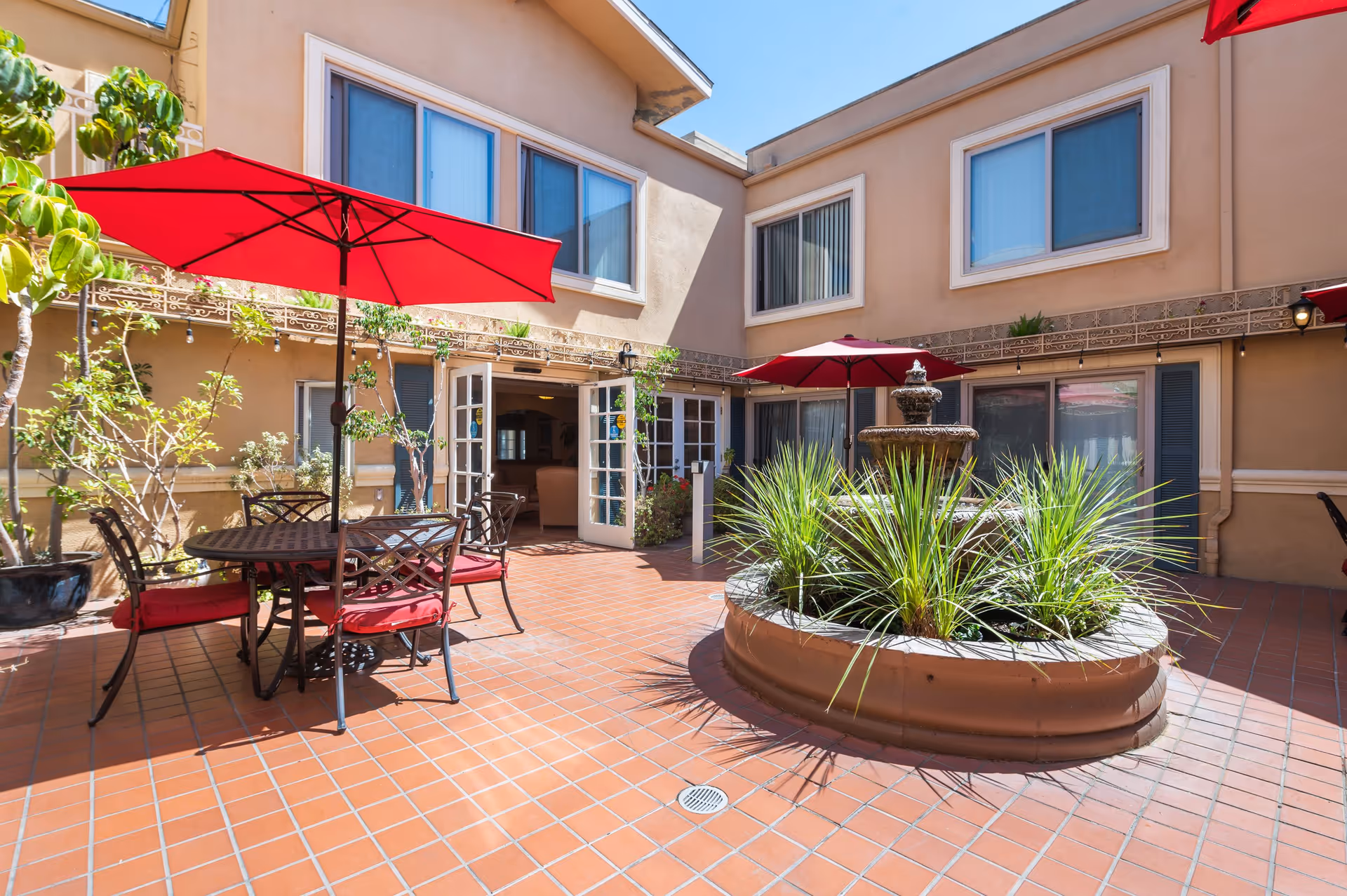 Outdoor courtyard area with terracotta tiled floor, round metal table and chairs with red cushions, and red umbrellas providing shade. A circular planter with green plants and a tiered water fountain is in the center. The courtyard is surrounded by a beige building with multiple windows and open French doors leading inside.