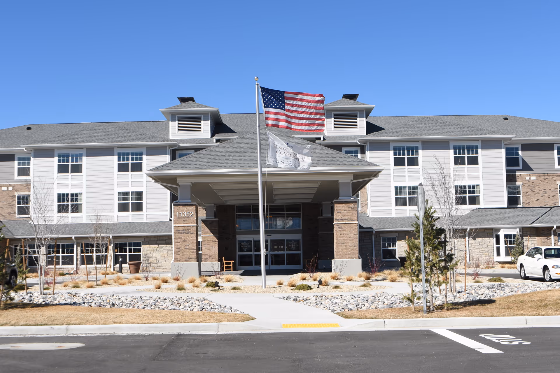 Front exterior view of South Jordan View Retirement Community building with an American flag and a community flag flying on flagpoles in front. The building has multiple windows, a covered entrance, and landscaping with rocks and small trees.