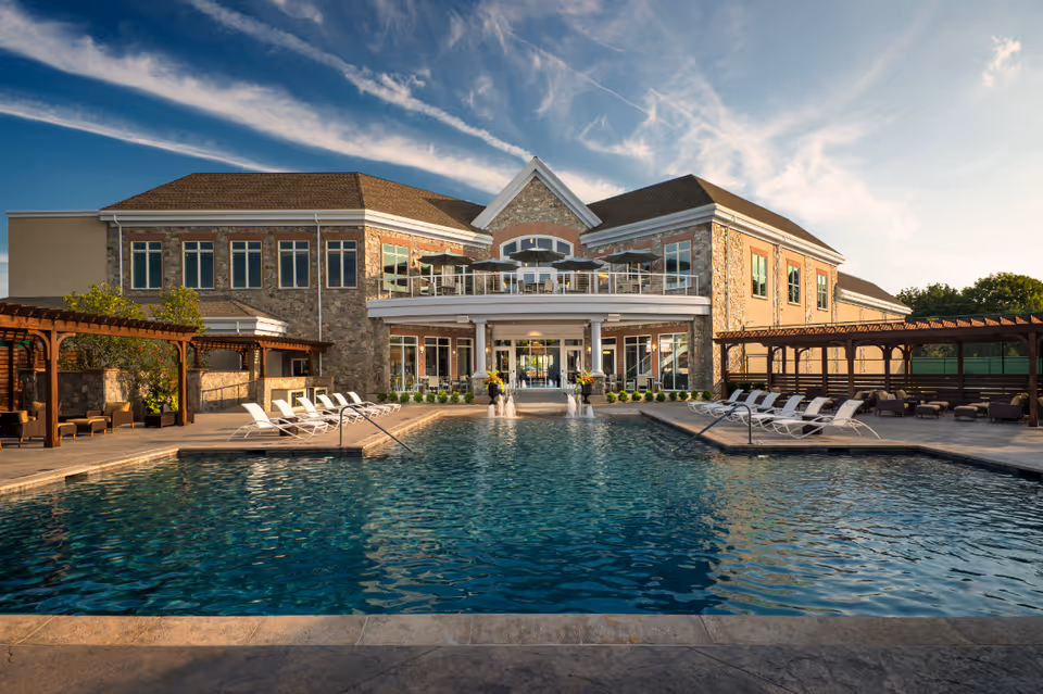 Outdoor swimming pool with lounge chairs on both sides, pergolas, and a large two-story stone building in the background under a partly cloudy sky.