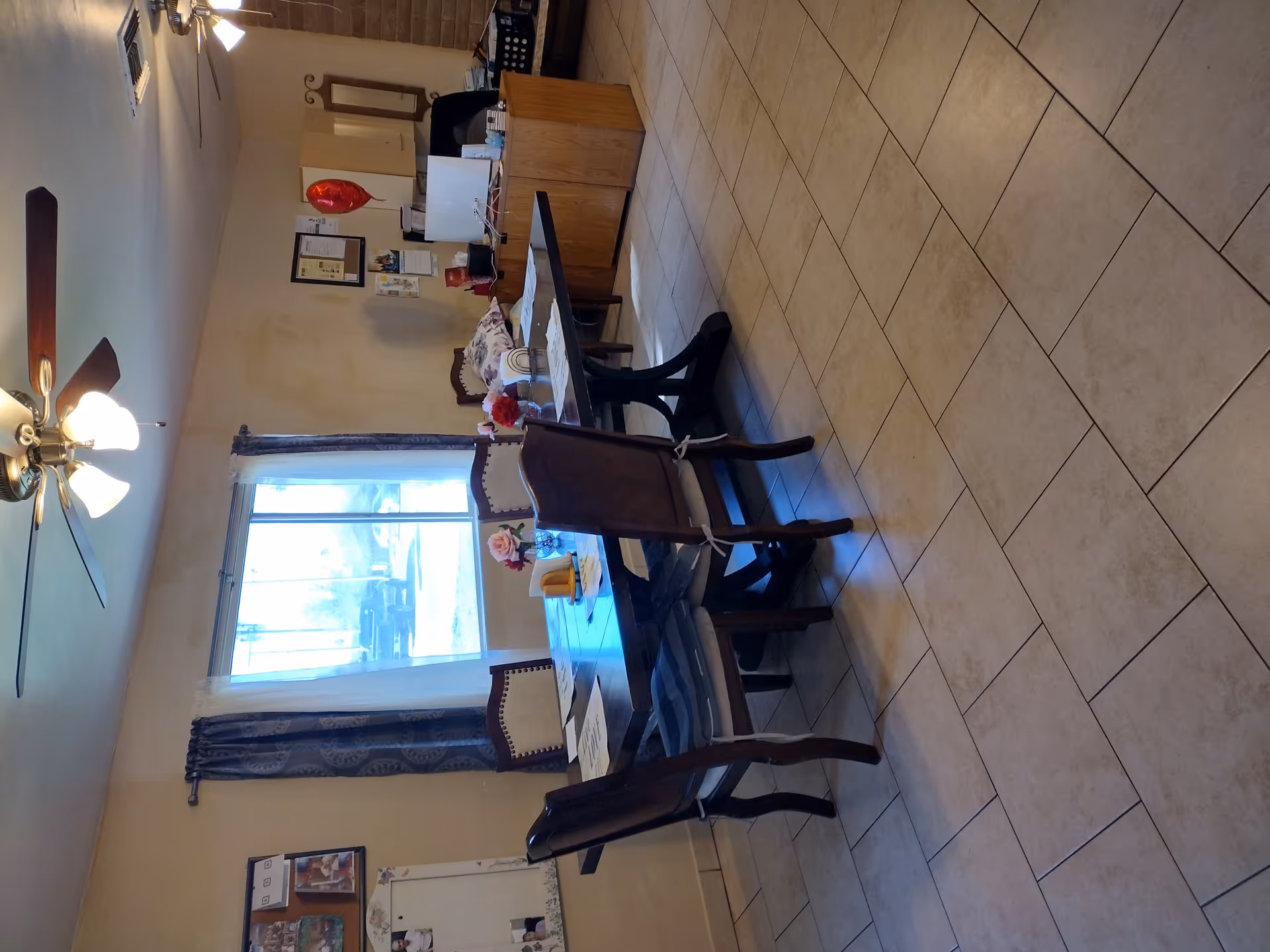 Interior view of a room with a rectangular dining table surrounded by six chairs. The table has placemats and small vases with flowers. There is a large window with curtains letting in natural light. In the background, there is a wooden desk with a computer monitor, papers, and a red heart-shaped balloon. The room has tiled flooring and ceiling fans with lights.