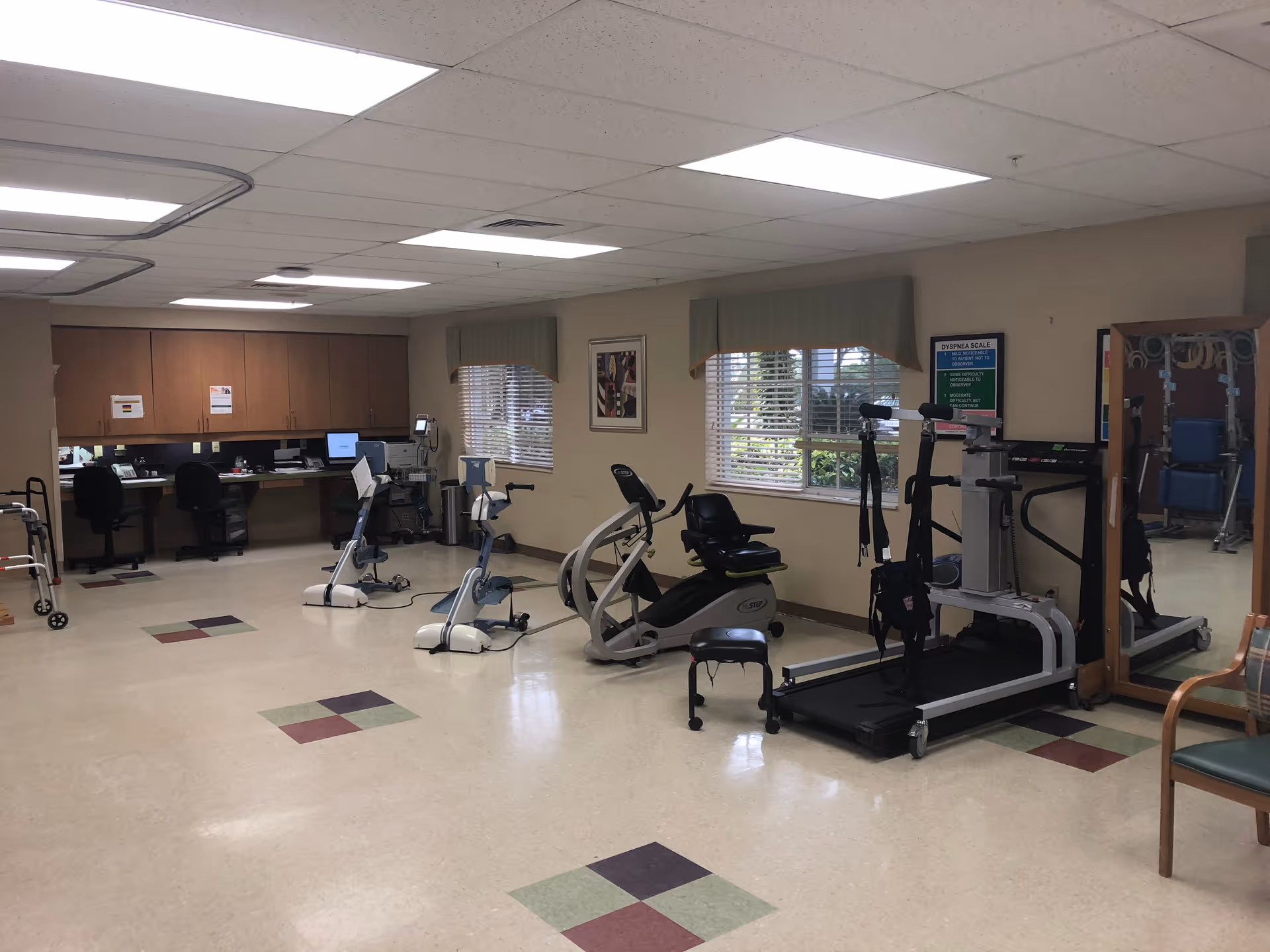 A physical therapy or rehabilitation room with exercise equipment including two stationary bikes, a recumbent stepper, and a treadmill with harness support. There are two windows with green valances, a large mirror on the right wall, and a desk area with two chairs and computer equipment on the left side of the room. The floor has a beige tile with colored square patterns.