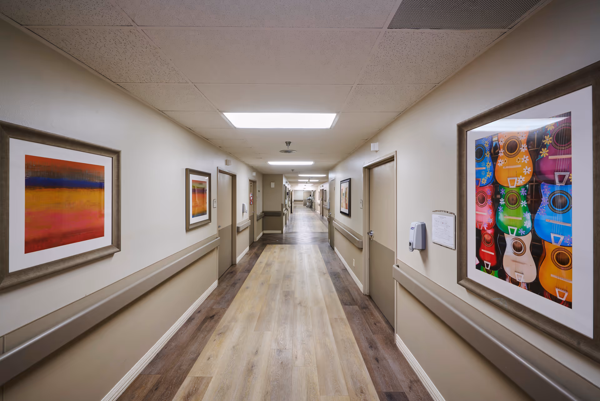 A long, well-lit hallway in a nursing center with light-colored walls and wood-patterned flooring. The walls are decorated with colorful framed artwork, including abstract paintings and images of guitars. Several closed doors line both sides of the hallway, and handrails run along the walls.