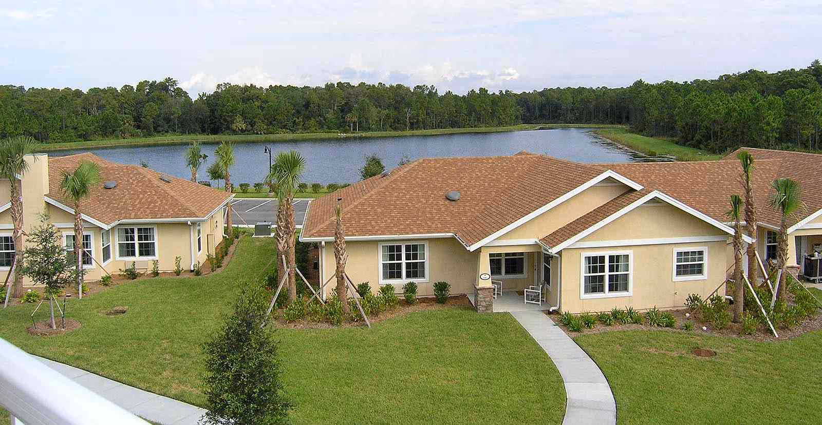 Single-story beige cottages with brown roofs, front lawns, palm trees and a lake in the background.