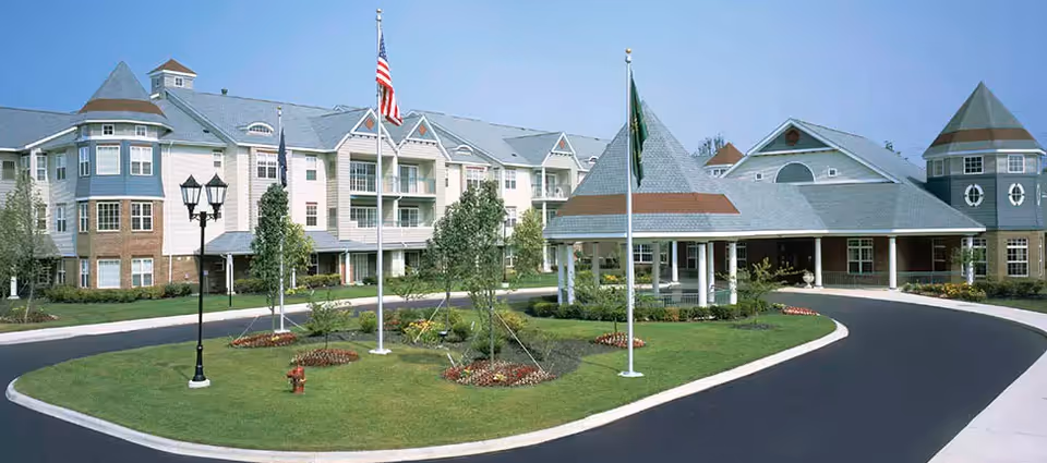 Exterior view of a large senior living facility building with multiple floors, featuring a covered entrance, landscaped lawn with trees and flower beds, and three flagpoles with flags in front of the building.