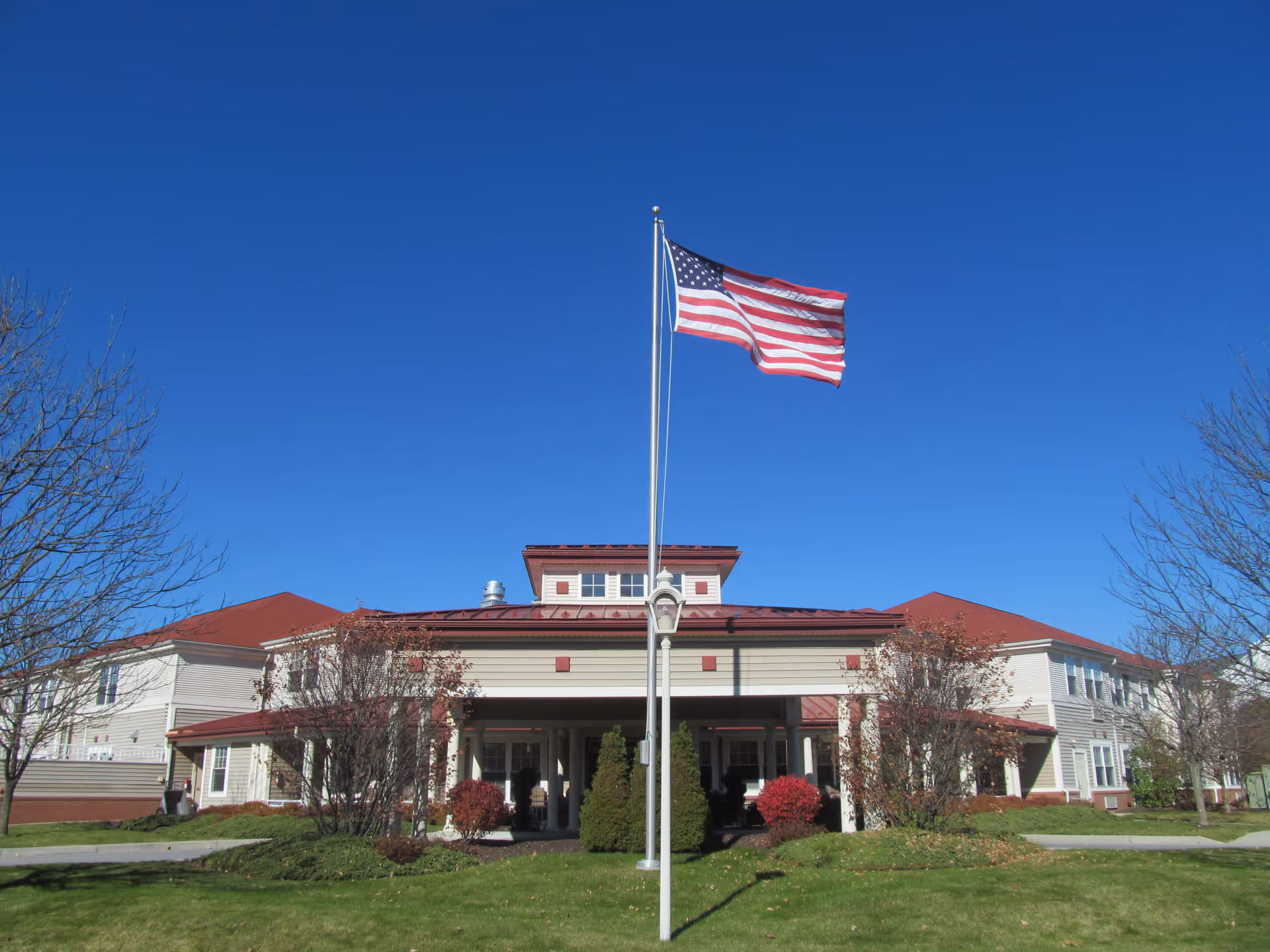 Exterior view of a senior living facility building with a red roof and beige siding under a clear blue sky. An American flag is flying on a flagpole in front of the building, surrounded by some trees and green grass.