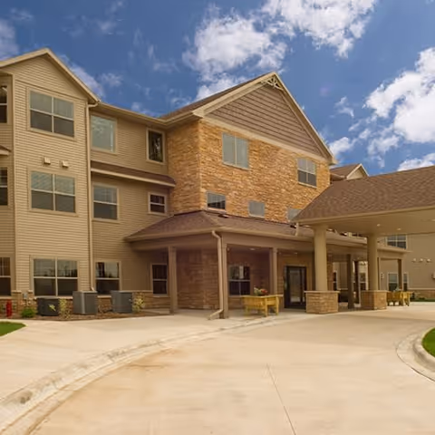 Exterior view of a multi-story senior living facility building with beige siding and stone accents under a partly cloudy blue sky. The entrance features a covered drop-off area supported by columns and a curved driveway.