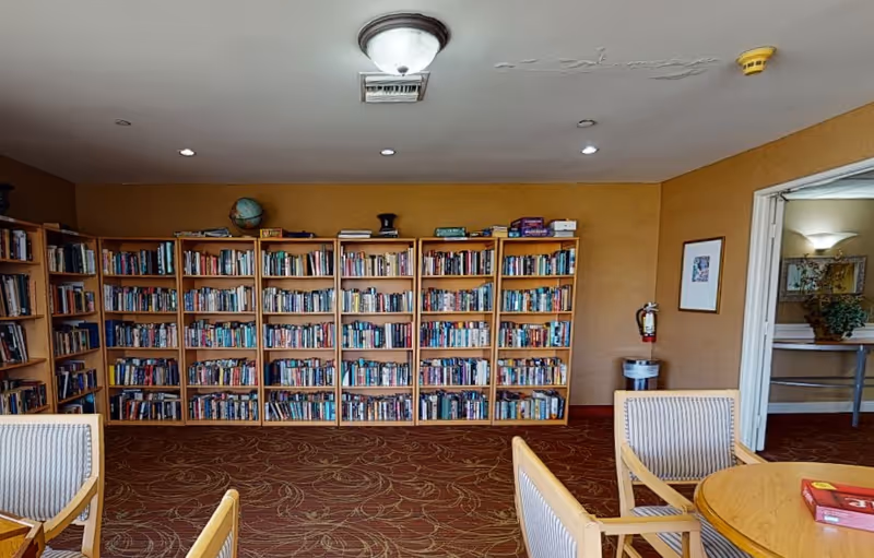 Interior room with multiple wooden bookshelves filled with books against a tan wall. There are several wooden chairs with striped cushions and a round wooden table with a board game on it. The ceiling has a light fixture and some visible damage. A fire extinguisher and a framed picture are on the right wall near an open doorway leading to another room with a plant and a wall sconce.