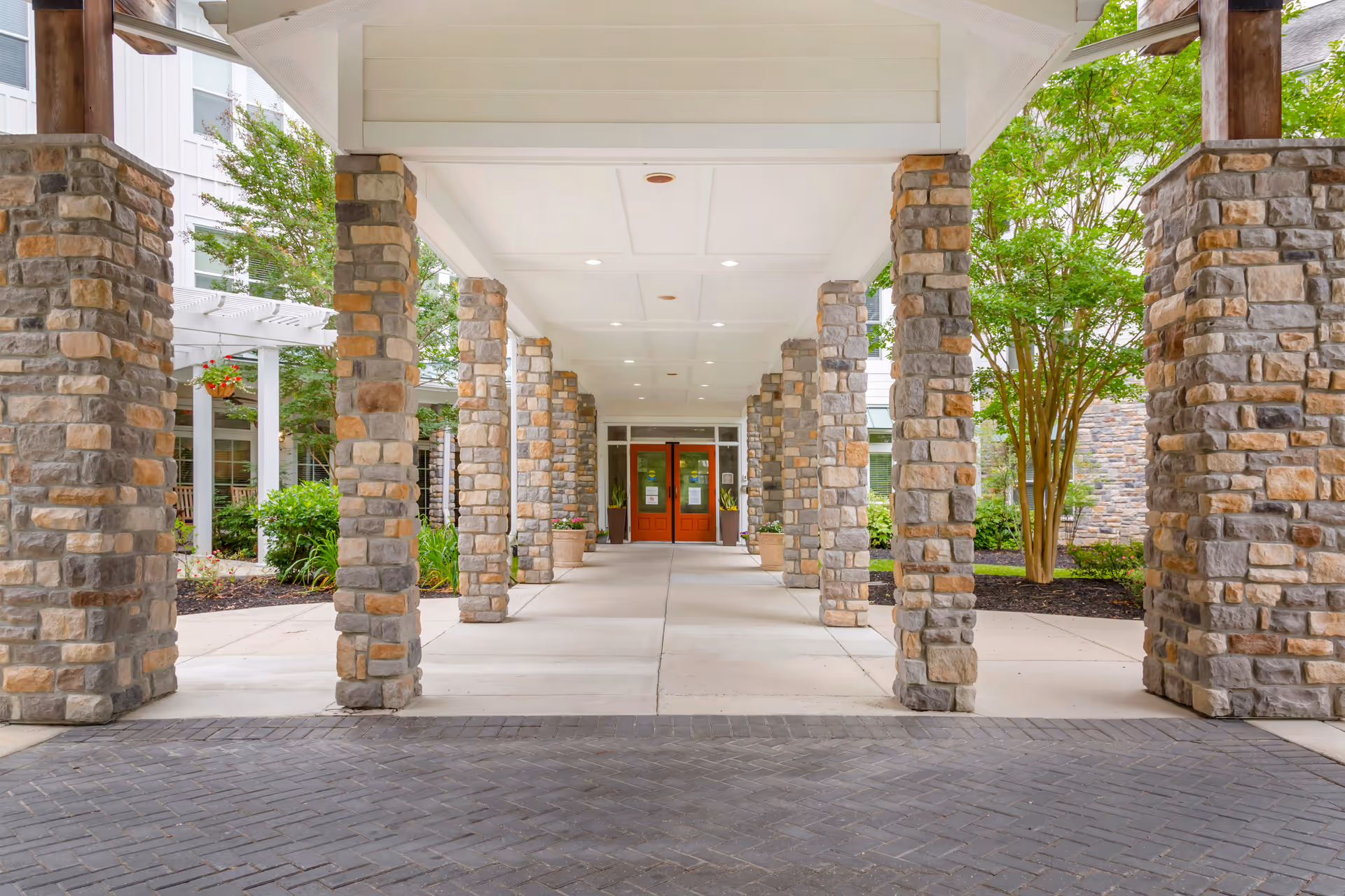 Covered entrance walkway supported by stone pillars leading to double wooden doors of a building, with greenery and trees on either side.