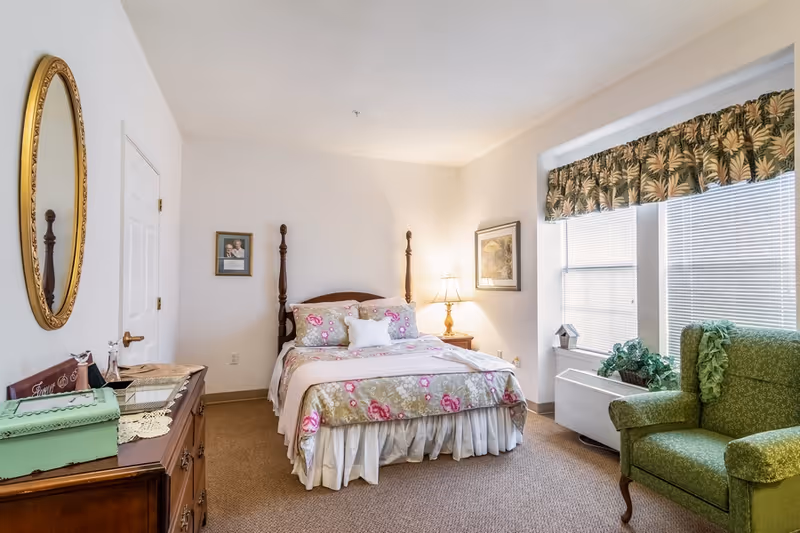 A cozy bedroom in a senior living facility with a wooden four-poster bed dressed in floral bedding, a wooden dresser with a green box and decorative items, a round gold-framed mirror on the wall, a nightstand with a lamp, framed pictures on the walls, a green upholstered armchair, and two windows with blinds and floral valances.
