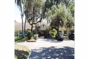 Front exterior view of a single-story residential building with a driveway and parked cars, surrounded by tall trees and greenery.