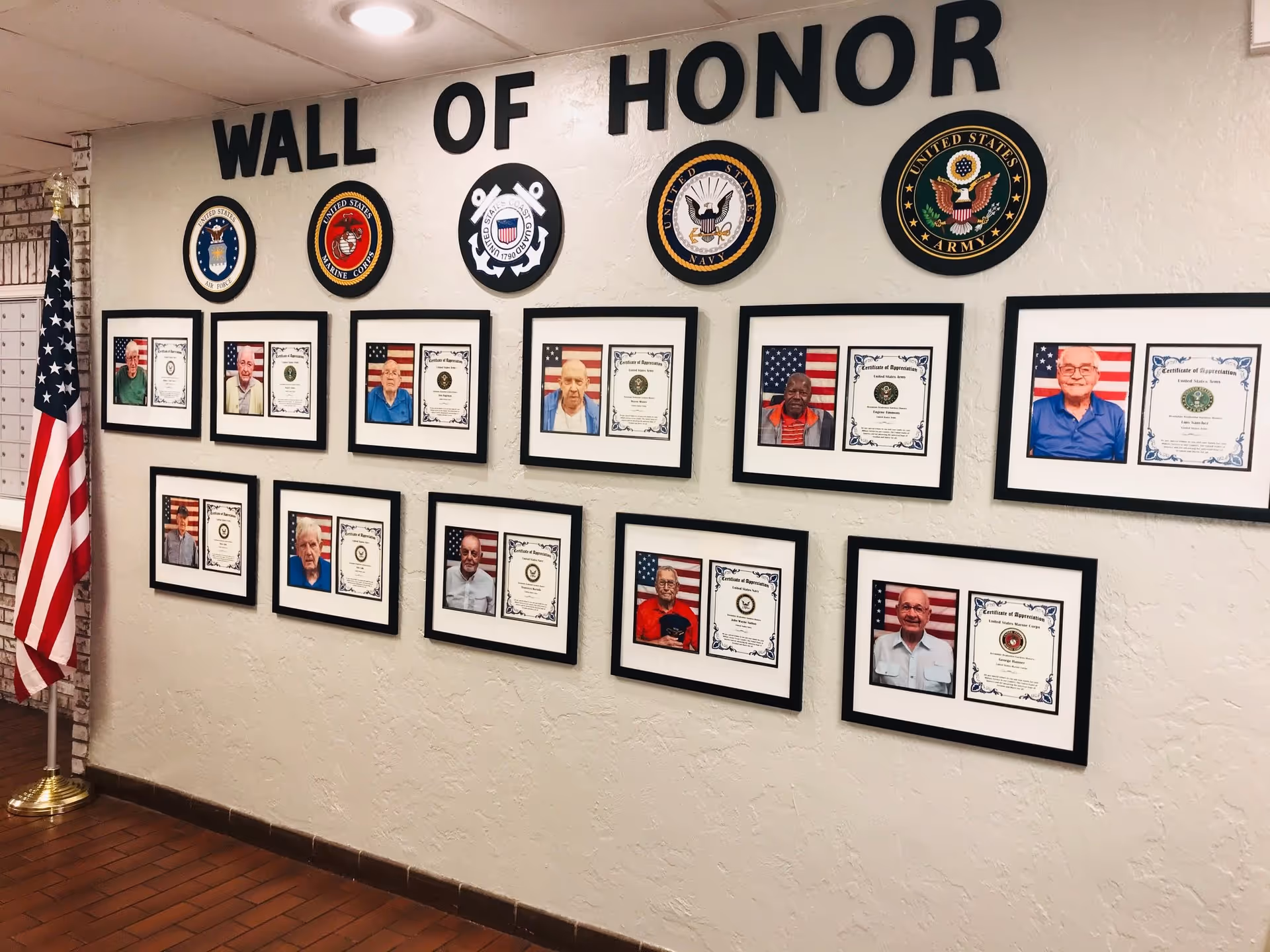 A wall display titled 'Wall of Honor' featuring framed photos and certificates of appreciation for veterans, with military branch emblems above and an American flag standing on a pole to the left.