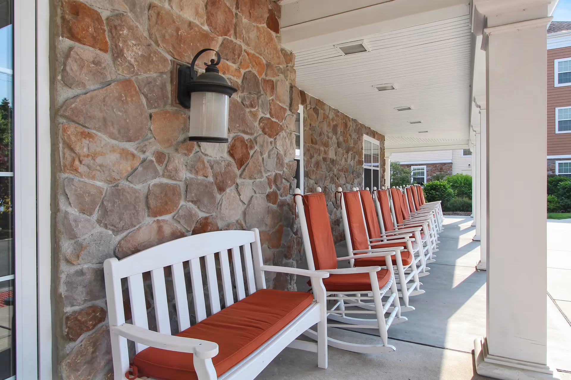 A row of white rocking chairs with orange cushions lined up on a covered porch with a stone wall and a white bench with an orange cushion in the foreground. There is a black outdoor wall lantern mounted on the stone wall.