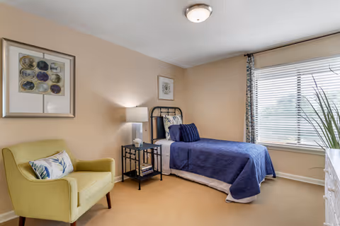 A cozy bedroom with a single bed covered in a blue quilt and white sheets. Next to the bed is a black metal nightstand with a white lamp and some books. A green upholstered armchair with a decorative pillow sits to the left. The room has beige walls, a large window with blinds and patterned curtains, and framed artwork on the walls.