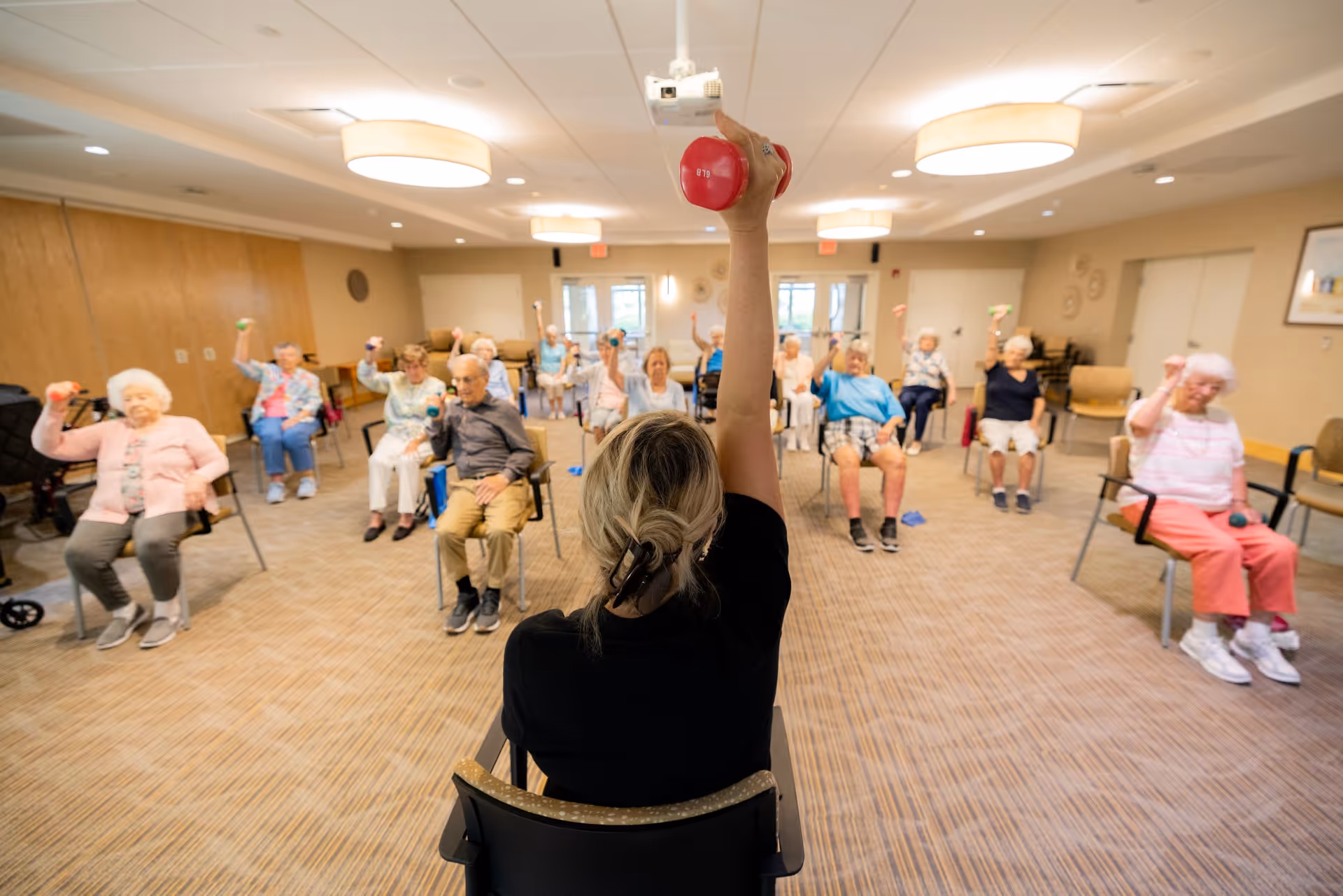 A group of elderly individuals seated in a spacious, well-lit room participating in a seated exercise class led by an instructor holding a red dumbbell. The participants are also holding small dumbbells and raising their arms in unison.