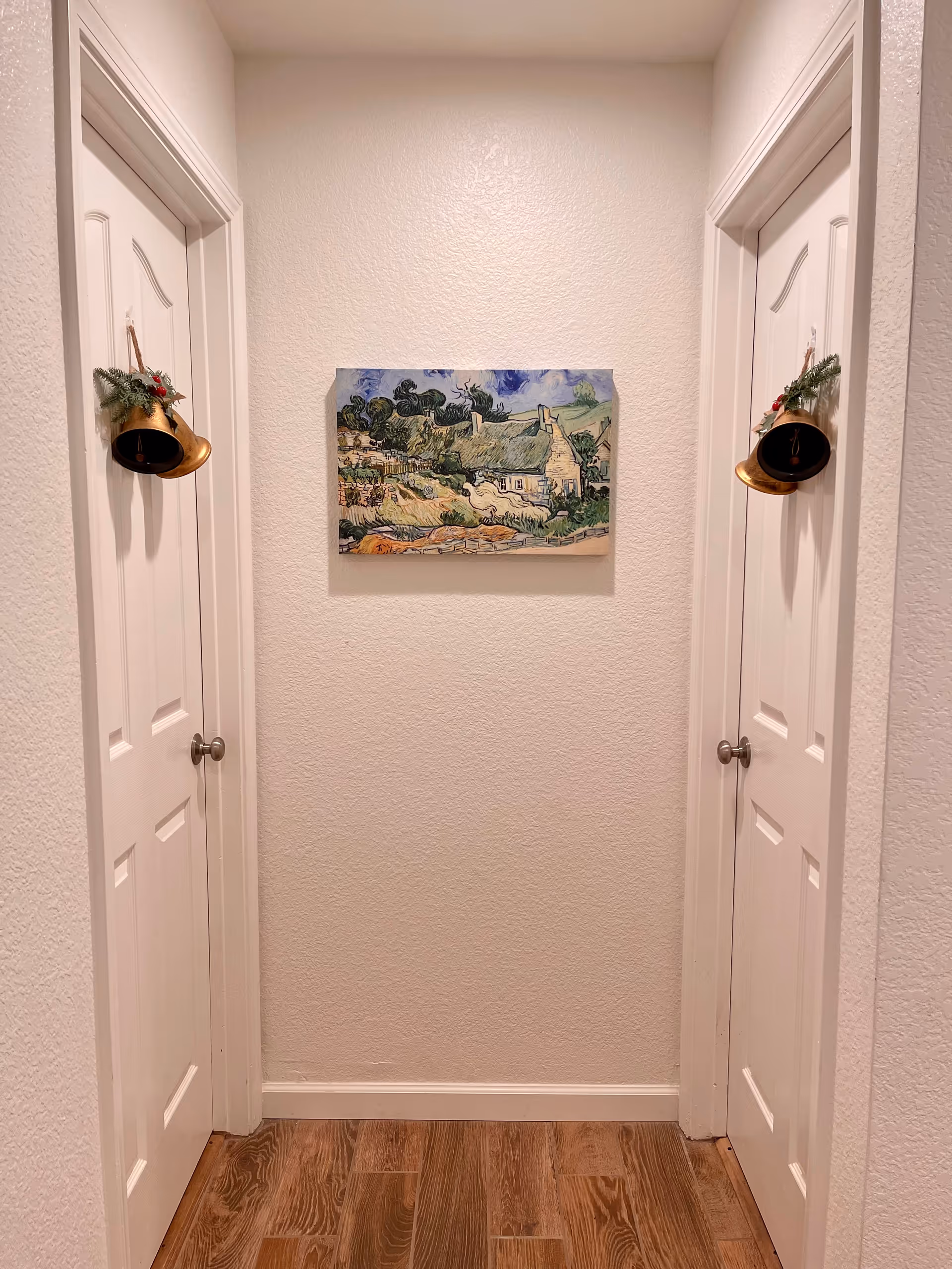 A narrow hallway with two white doors facing each other, each decorated with a hanging golden bell adorned with greenery and red berries. A colorful painting of a countryside scene is centered on the wall between the doors. The floor is wooden with a warm tone, and the walls are painted light beige.