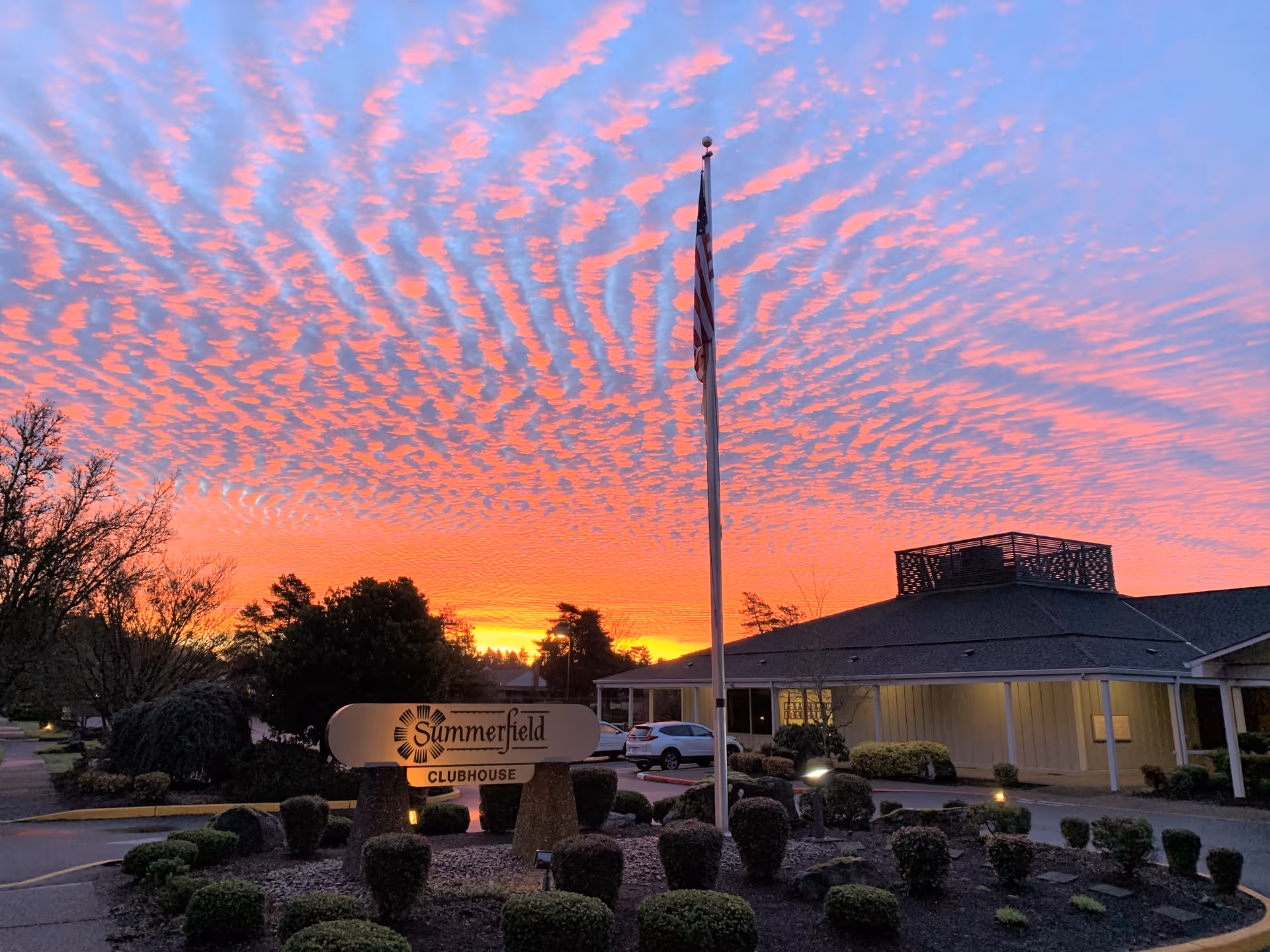 Sunset sky with vibrant orange and pink clouds over the Summerfield Civic Association clubhouse building, with a flagpole and landscaped bushes in the foreground.