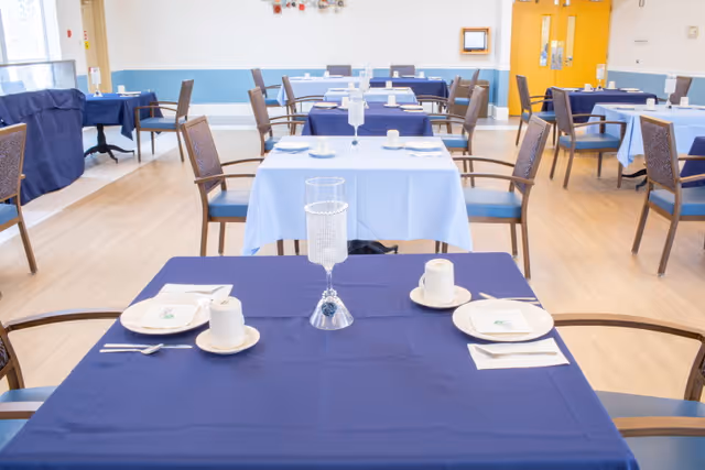 Dining room with multiple tables covered in blue tablecloths set with plates, cups and chairs in a senior facility.