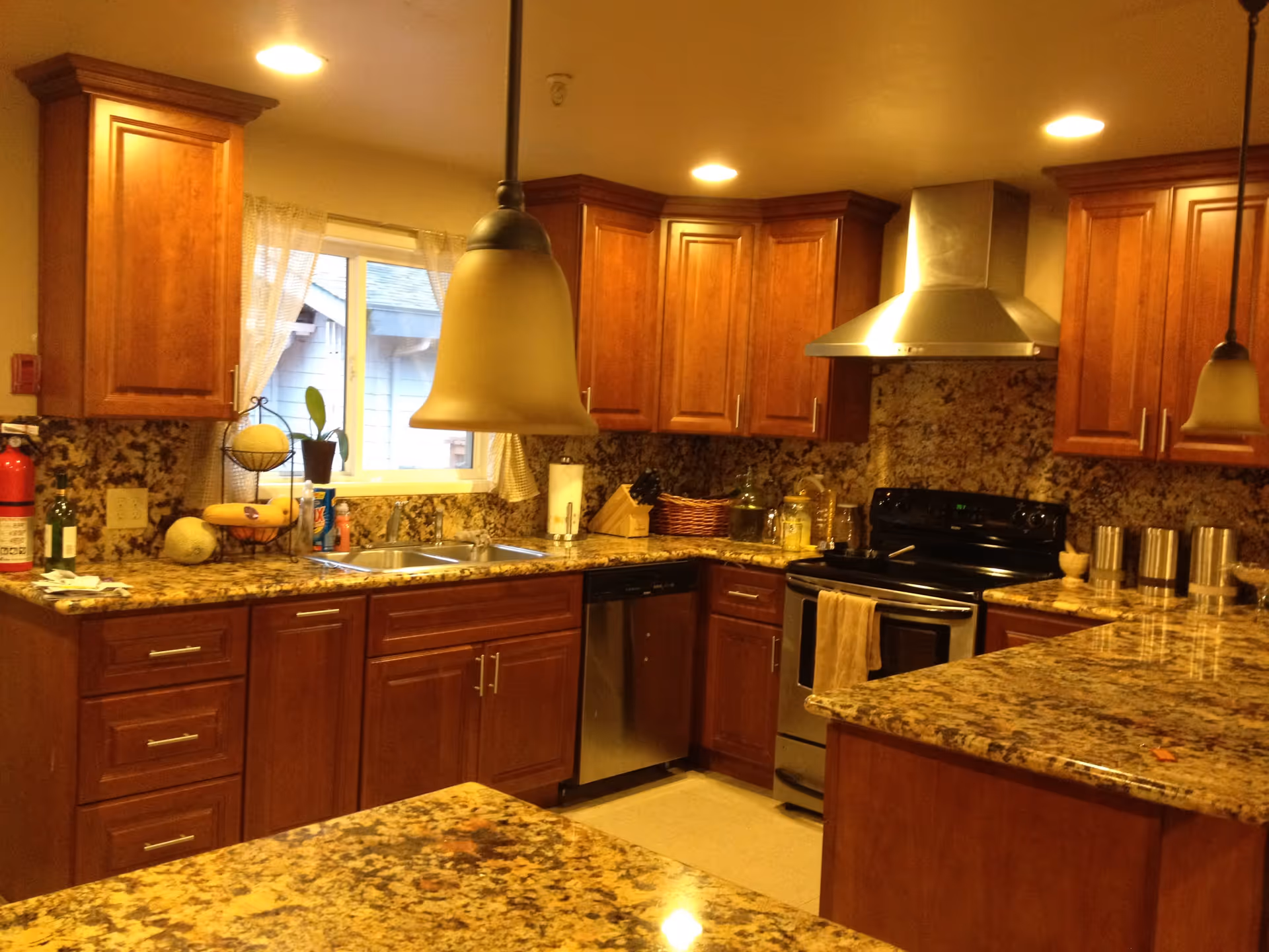 A warm-toned kitchen with wooden cabinets, granite countertops, and a stainless steel stove and dishwasher. There are two pendant lights hanging from the ceiling, a window with sheer curtains above the sink, and various kitchen items such as a fire extinguisher, fruit basket, and containers on the counters.