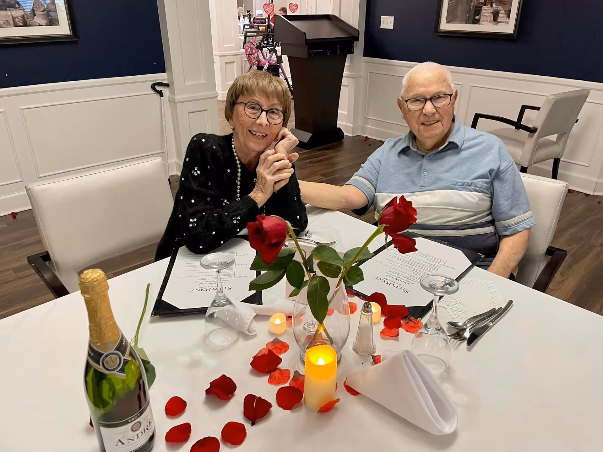 An elderly couple sitting at a dining table in a decorated room, holding hands and smiling. The table is set with menus, wine glasses, a bottle of champagne, red rose petals, a vase with red roses, and a lit candle, creating a romantic atmosphere.