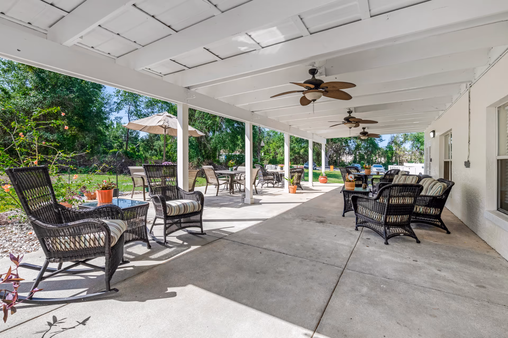 Covered outdoor patio area with multiple wicker chairs and tables, ceiling fans, and potted plants. The patio overlooks a green garden with trees and an umbrella-covered table in the background.