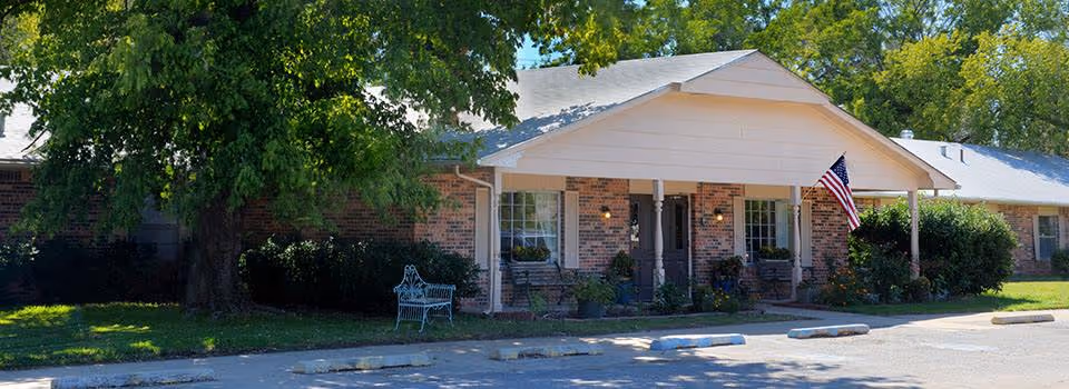 Exterior view of a single-story brick building with a covered entrance supported by columns, an American flag mounted near the door, surrounded by trees and bushes, and a parking area with marked spaces in front.