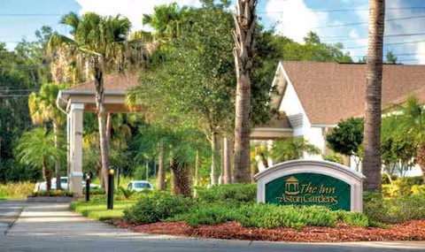 Exterior view of Aston Gardens senior living facility entrance with landscaping and signage.