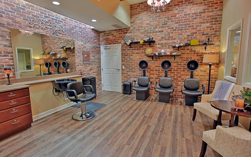 Interior view of a salon area with three black hair dryer chairs against a brick wall, a single styling chair in front of a large mirror, wooden flooring, and two beige patterned chairs with a small round table between them.