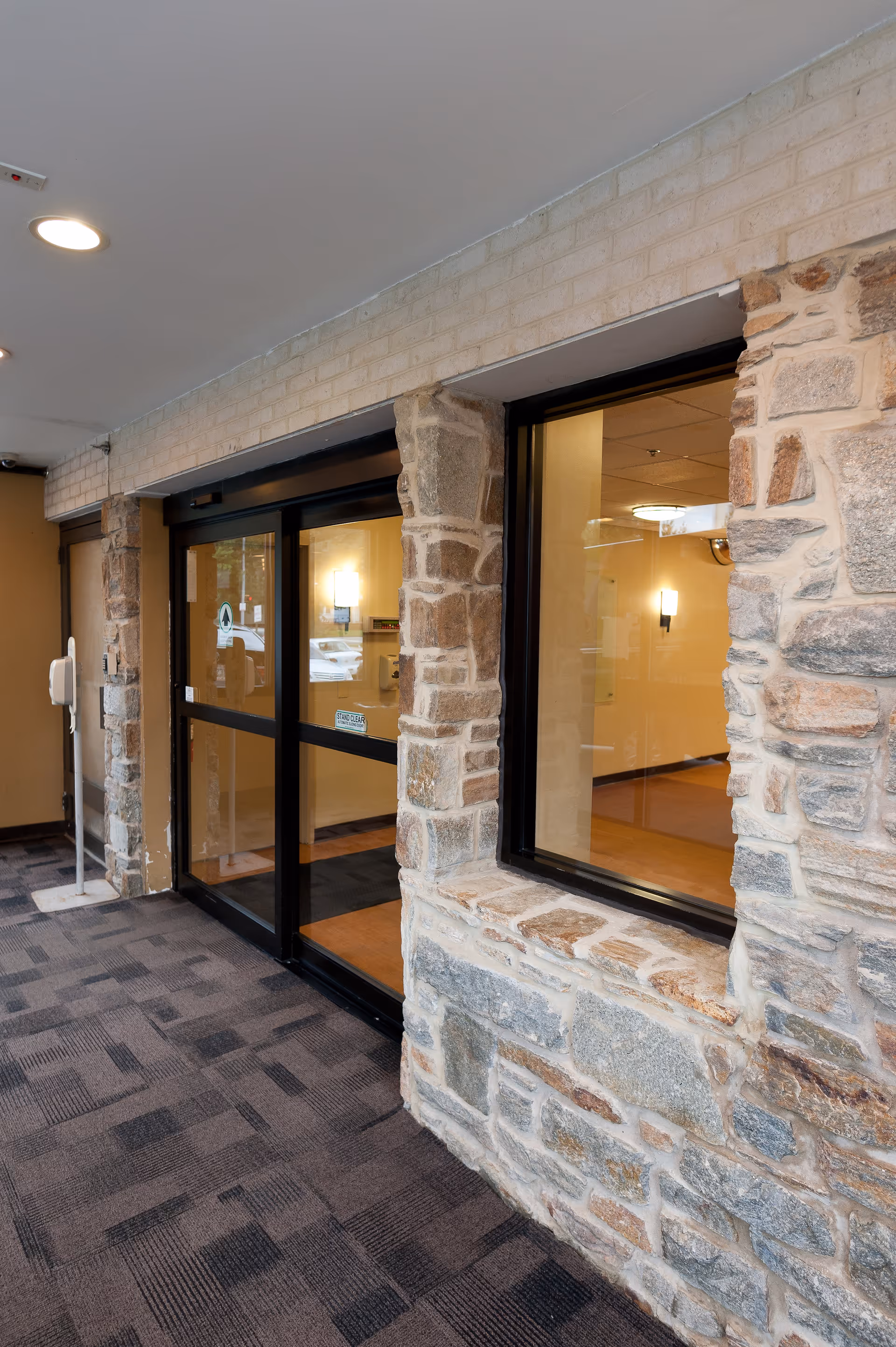 Entrance area of a facility with stone pillars and walls, featuring automatic sliding glass doors and a large window. The floor is carpeted with a patterned design, and the interior visible through the glass has warm lighting and beige walls.