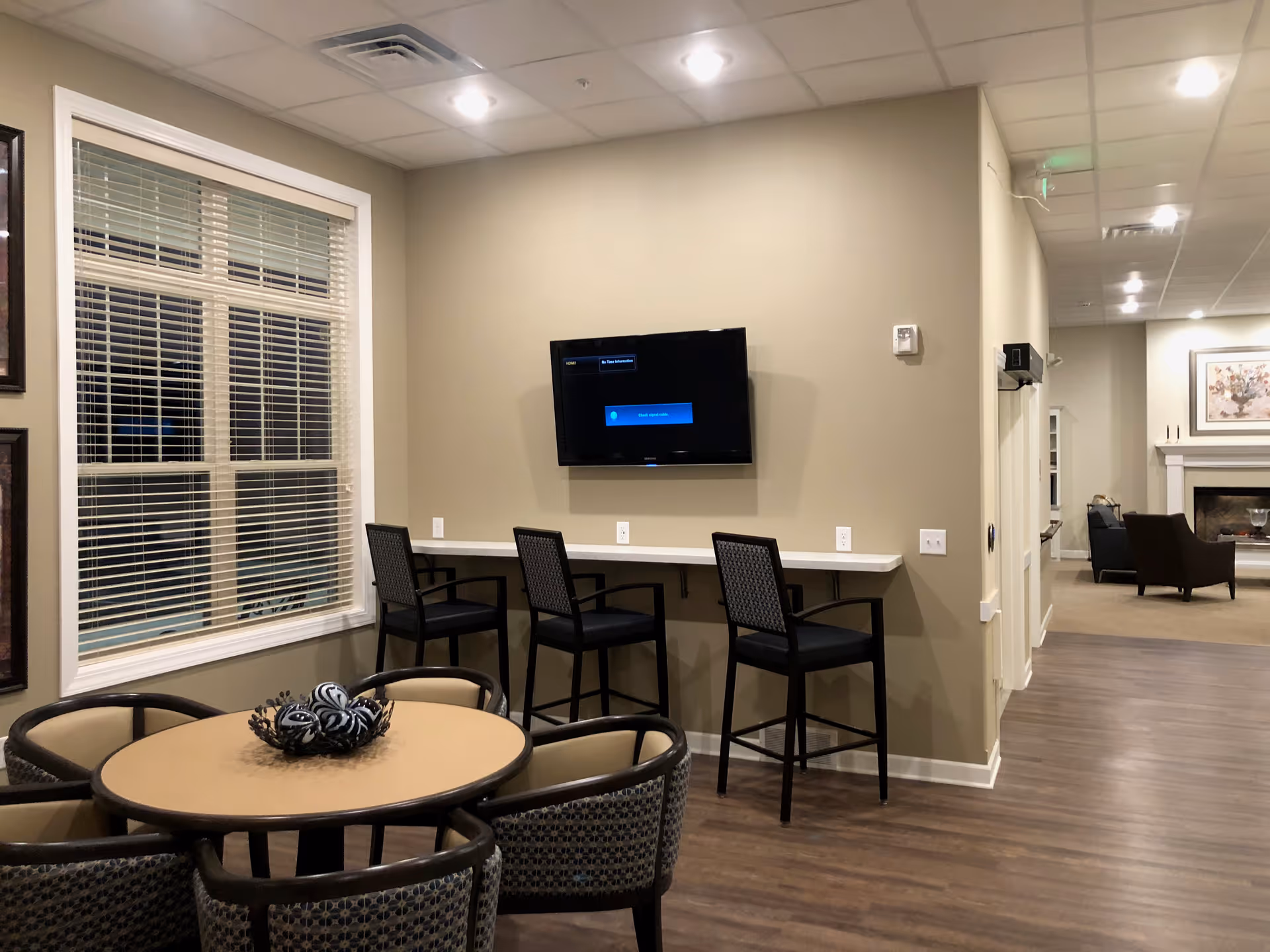 A senior living common area with a round table and chairs, a wall-mounted TV above a counter with three bar stools, and seating with a fireplace visible in the background.