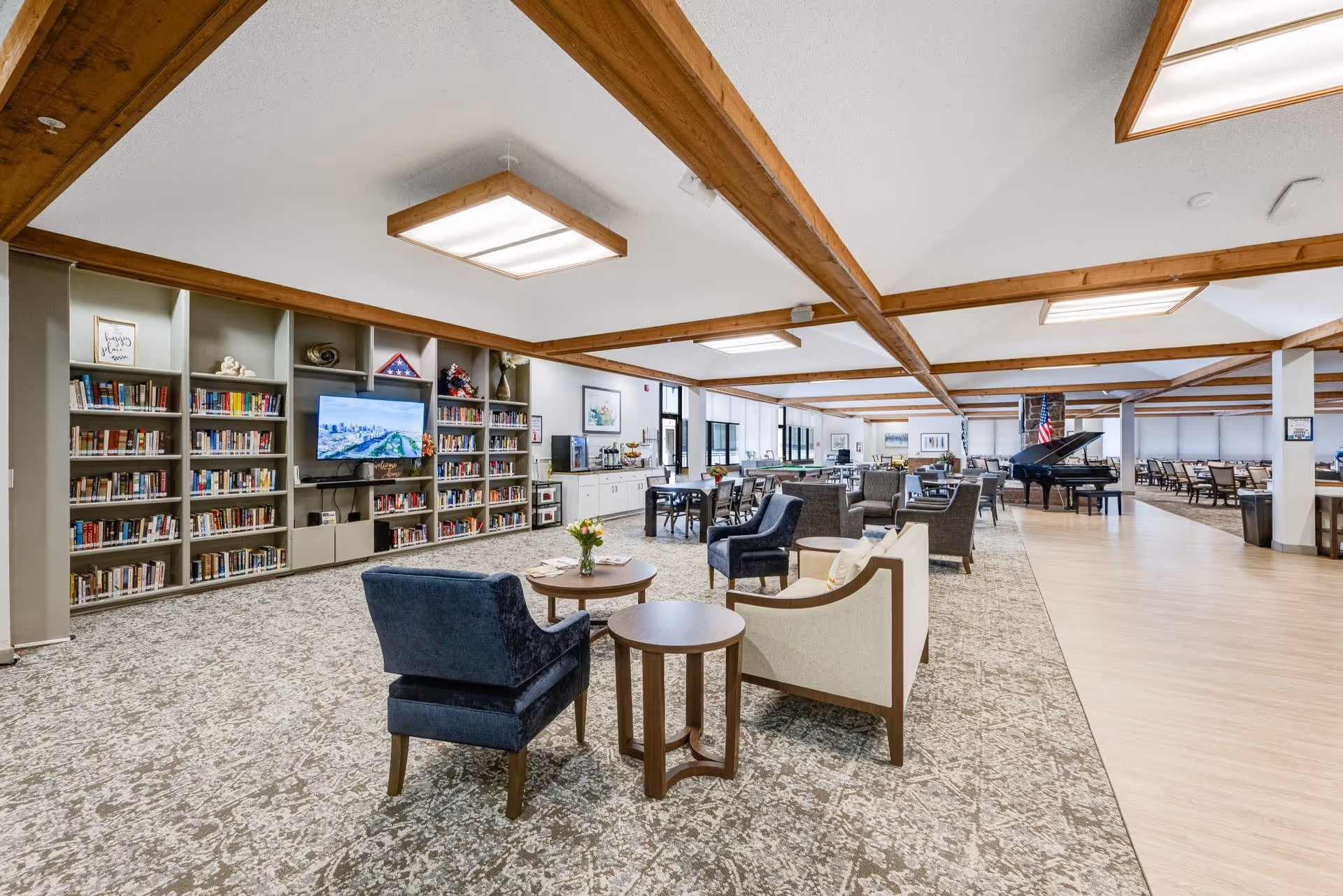 Spacious senior living common area with bookshelves, seating clusters, and a grand piano under exposed wood beams.