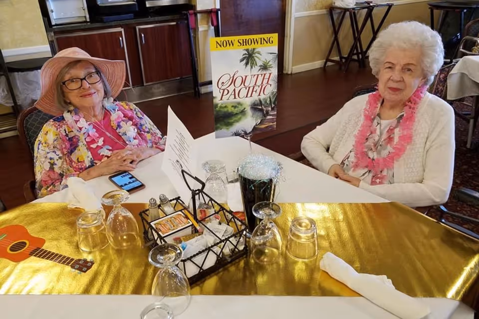 Two elderly women wearing leis seated at a decorated dining table with a 'Now Showing South Pacific' sign.