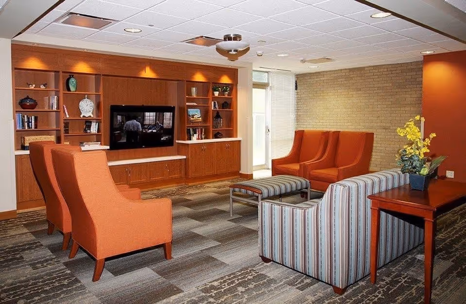 A furnished lounge with orange armchairs, a striped sofa and ottomans facing a TV set in wooden built-in shelving, with a side table holding yellow flowers.