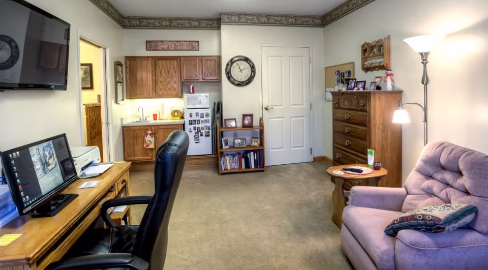 Interior view of a senior living facility room featuring a small kitchenette with wooden cabinets, a microwave, and a refrigerator. A wall clock hangs above a small bookshelf with framed photos and books. To the right, there is a wooden dresser with more framed photos and a floor lamp next to a purple upholstered armchair with a decorative pillow. On the left side, there is a wooden desk with a computer monitor, printer, and a black office chair. The room has beige carpet and light-colored walls with decorative crown molding.