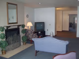 A small common room with a fireplace, mirror, TV, upholstered chairs and a couch facing a hallway.